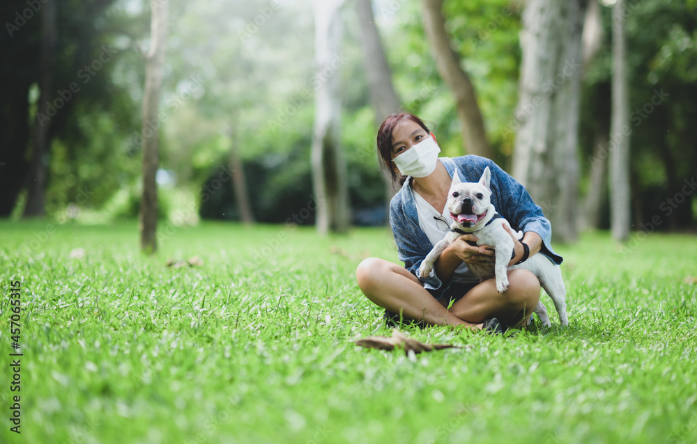 Fototapeta premium A woman wearing a medical mask sitting on the lawn with a white French bulldog.