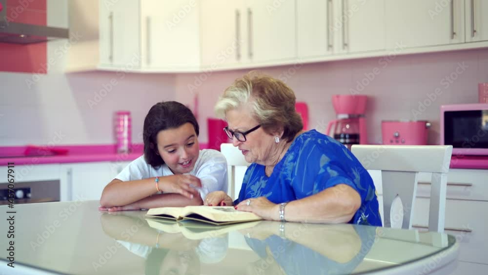 Elderly woman reading book with granddaughter in light kitchen at home Elderly woman reading book with granddaughter in light kitchen at home
