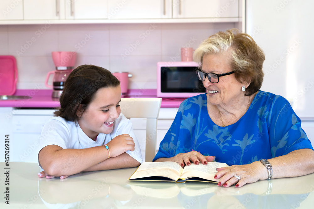 Elderly woman reading book with granddaughter in light kitchen at home Elderly woman reading book with granddaughter in light kitchen at home