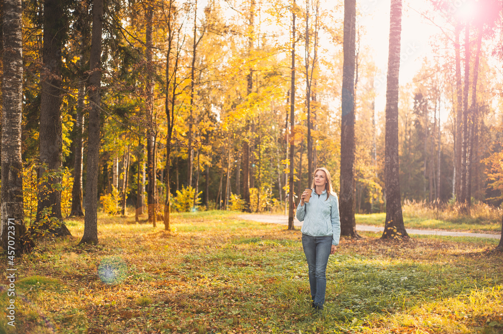 A beautiful blonde woman walks through the autumn park with a cup of warming drink in her hands. Walk on a sunny autumn evening. Healthy lifestyle.