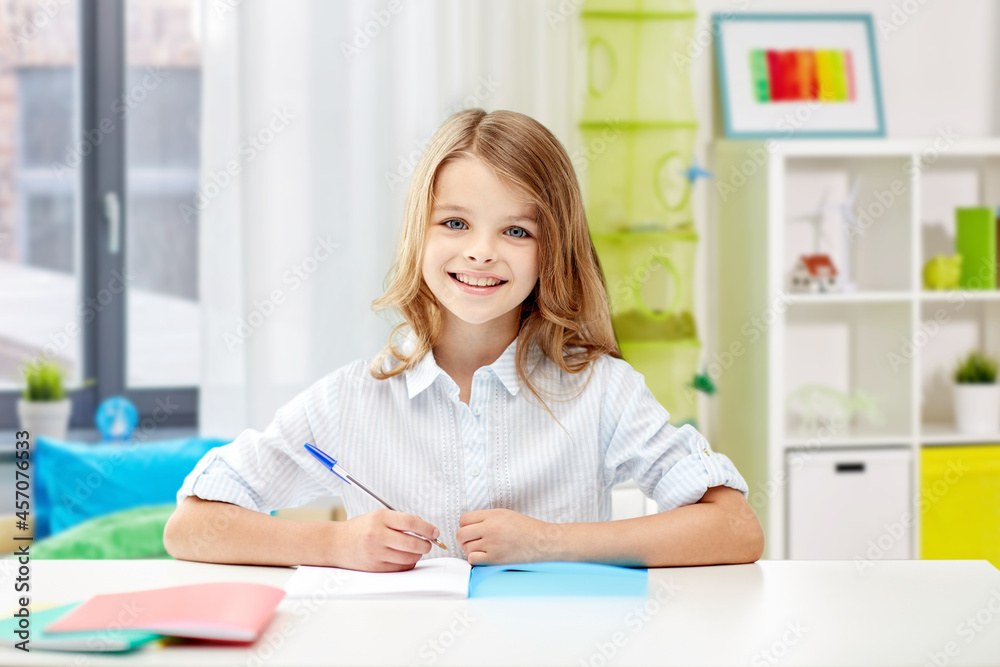 education, elementary school, learning and people concept - happy smiling girl with notebook and pen sitting at table over home background
