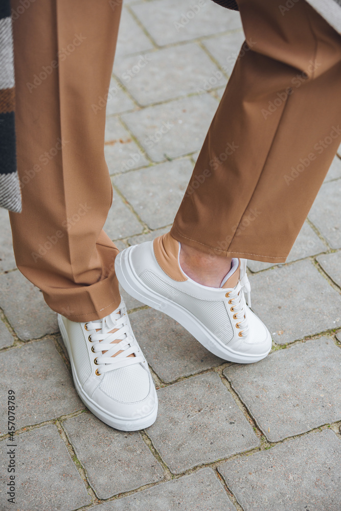 Closeup photo of beautiful fashionable woman wearing checkered long coat, beige pants, white blouse and sneakers.