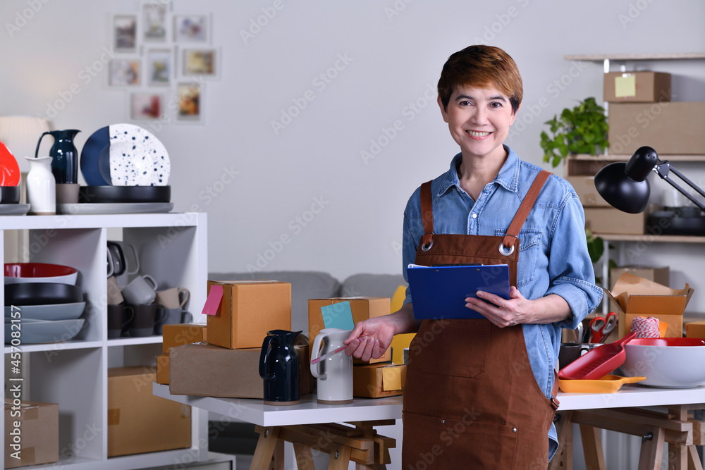 Fototapeta premium Mature Asian woman entrepreneur, Business owner standing in front of her clay ceramic product and working at home