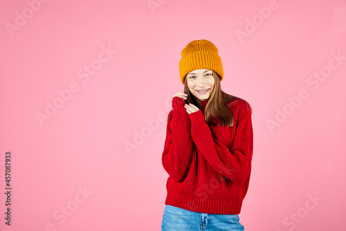 Happy young woman wearing warm sweater and knitted hat on pink background. Girl winter portrait. Smiling hipster with braces having fun.