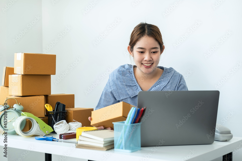 Asian woman small business owner holding box of parcel in hands and checking order on laptop while courier working Asian woman small business owner holding box of parcel in hands and checking order on laptop while courier working