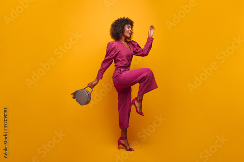 full-length stunning feminine lady waving her hands raising her leg to knee isolated in photo studio. young woman dressed in lilac loose suit and open high-heeled shoes.