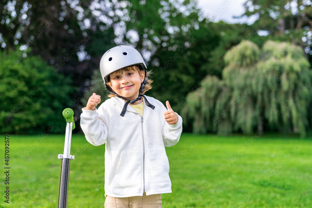 Happy little boy wearing safety helmet staying near kick scooter . Close up portrait of a smiling child walking in a park. Happy little boy wearing safety helmet staying near kick scooter . Close up portrait of a smiling child walking in a park.
