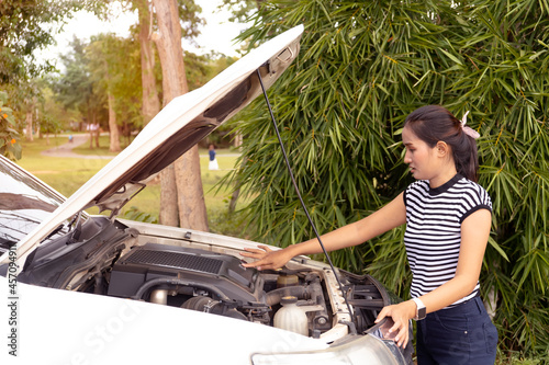Female driver standing on street near her car with popped up hood looking at broken engine.