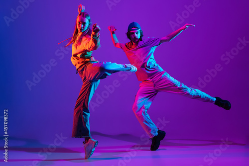 Fototapeta Naklejka Na Ścianę i Meble -  Two dancers, young man and woman dancing hip-hop in casual sports youth clothes on gradient purple pink background at dance hall in neon light.