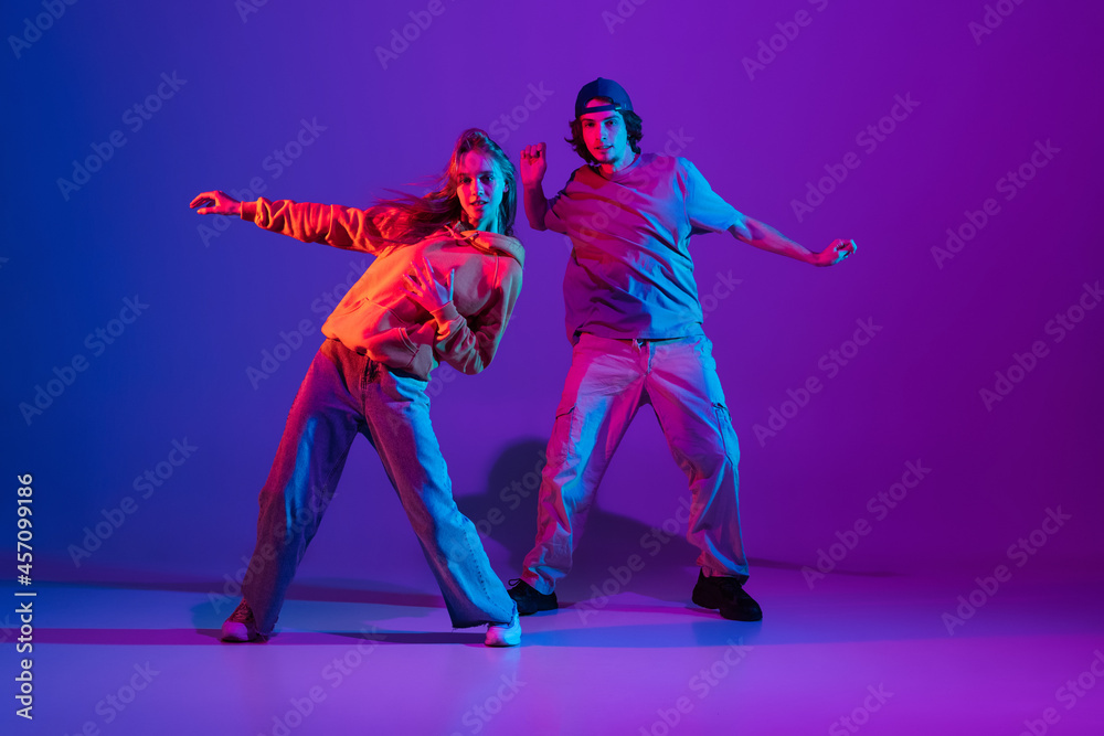 Young man and woman dancing hip-hop in casual sports youth clothes on gradient purple pink background at dance hall in neon light.