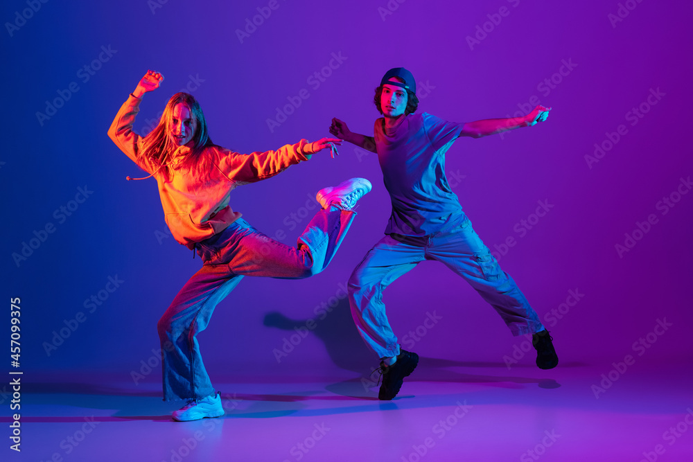 Young man and woman dancing hip-hop in casual sports youth clothes on gradient purple pink background at dance hall in neon light.