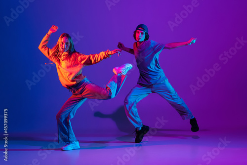 Fototapeta Naklejka Na Ścianę i Meble -  Young man and woman dancing hip-hop in casual sports youth clothes on gradient purple pink background at dance hall in neon light.