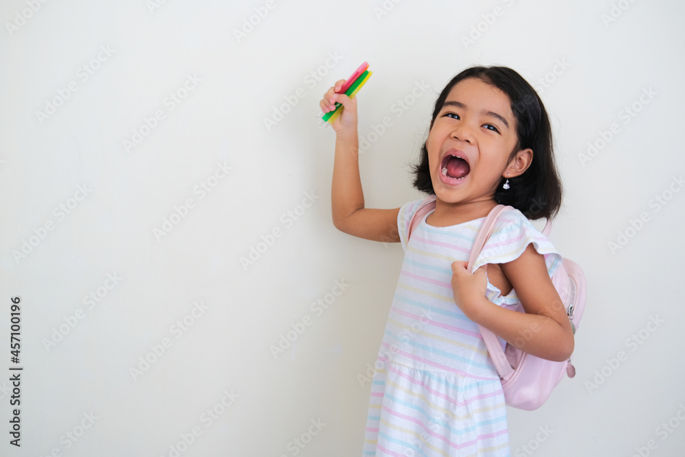 Asian little girl showing excited face expression while wearing backpack and holding coloring pen Asian little girl showing excited face expression while wearing backpack and holding coloring pen