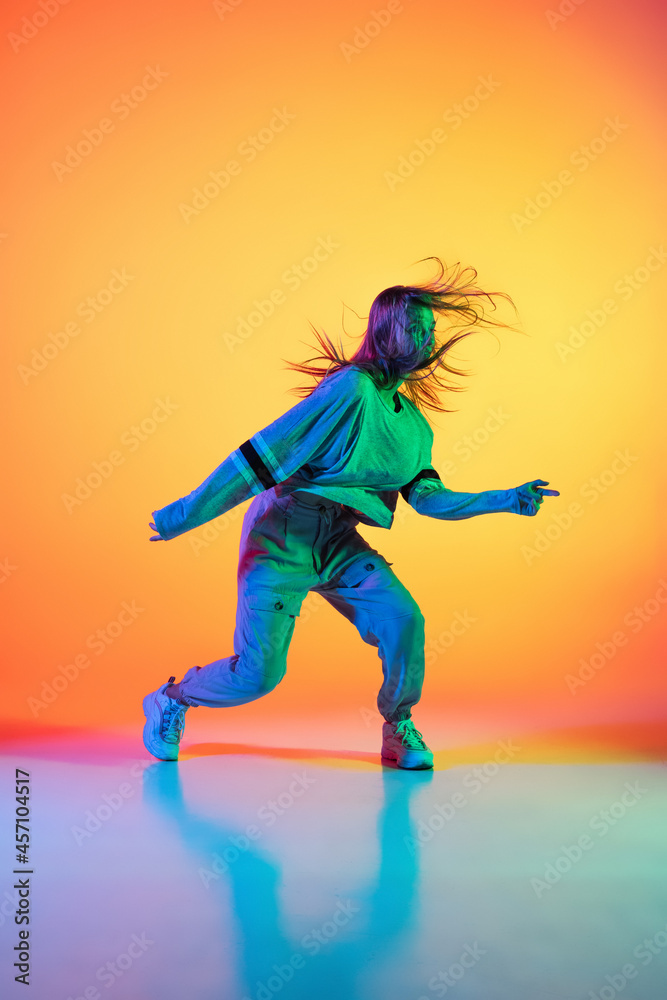 Young sportive girl dancing hip-hop in stylish clothes on colorful background at dance hall in neon light. Youth culture, movement, style and fashion, action.