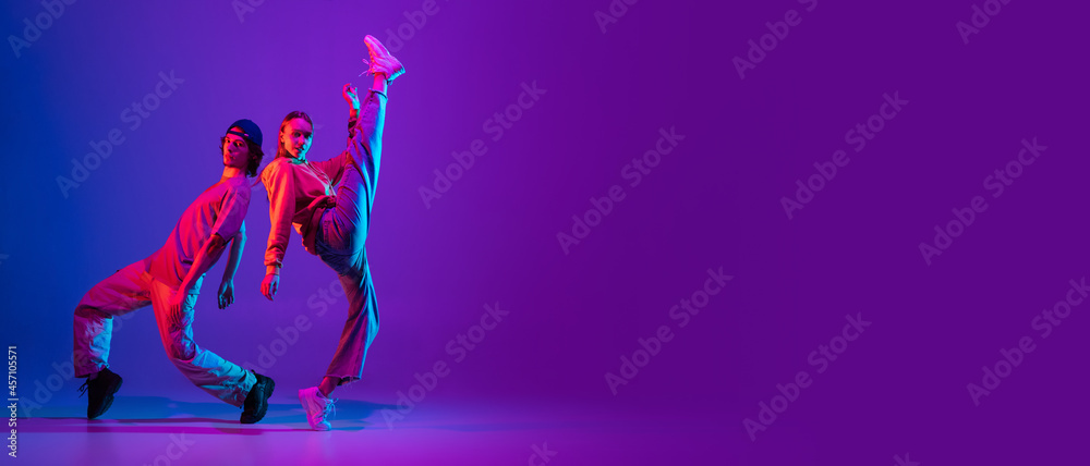Fototapeta premium Flyer. Two dancers, young man and woman dancing hip-hop in casual sports youth clothes on gradient purple pink background at dance hall in neon light.