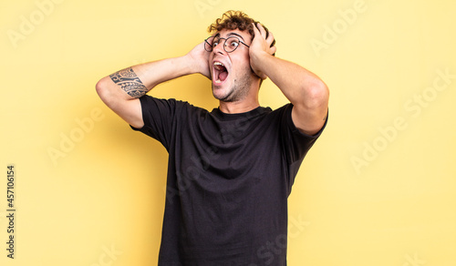 young handsome man with open mouth, looking horrified and shocked because of a terrible mistake, raising hands to head