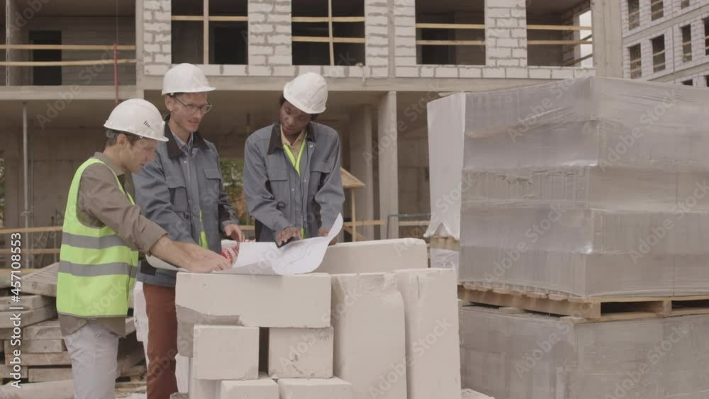 Medium long of three multiethnic architects wearing work outfits with hard hats, looking at paper with blueprint, having discussion in foreground of multistory building under construction Medium long of three multiethnic architects wearing work outfits with hard hats, looking at paper with blueprint, having discussion in foreground of multistory building under construction