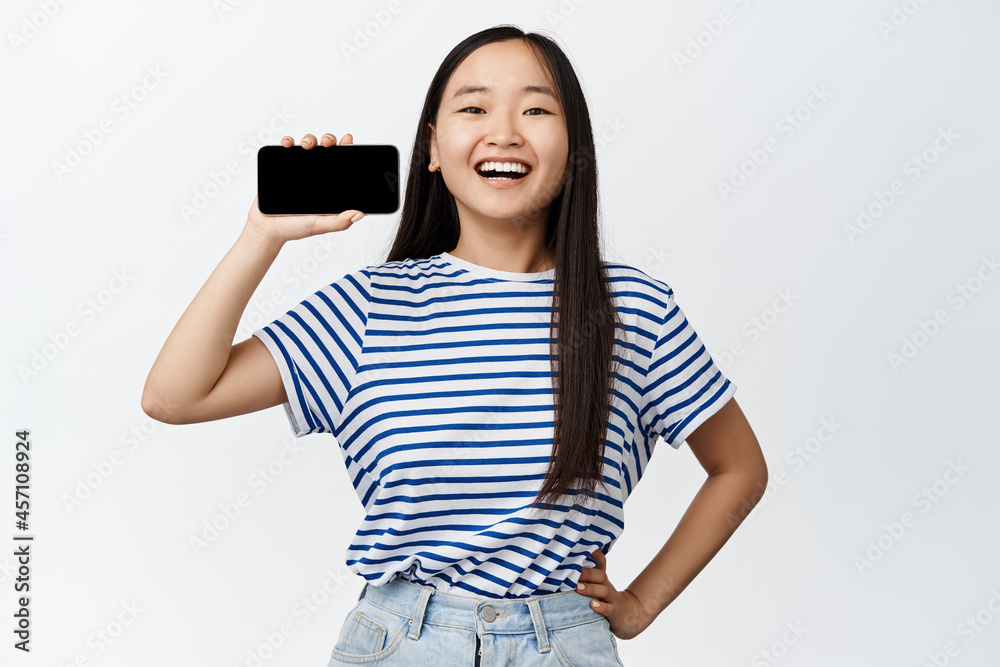 Portrait of excited asian girl shows her mobile phone horizontal screen, smiling pleased, recommending application, white background Portrait of excited asian girl shows her mobile phone horizontal screen, smiling pleased, recommending application, white background