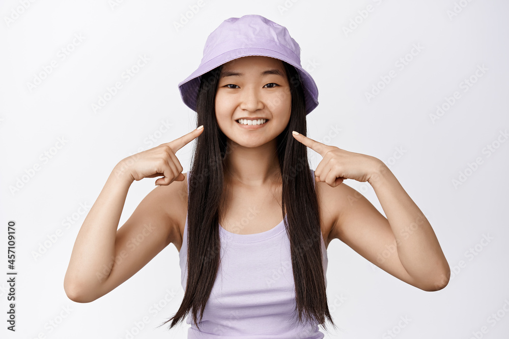 Portrait of asian young woman in bucket hat, pointing fingers at her white smile, concept of dental clinic and teeth whitening, studio background Portrait of asian young woman in bucket hat, pointing fingers at her white smile, concept of dental clinic and teeth whitening, studio background