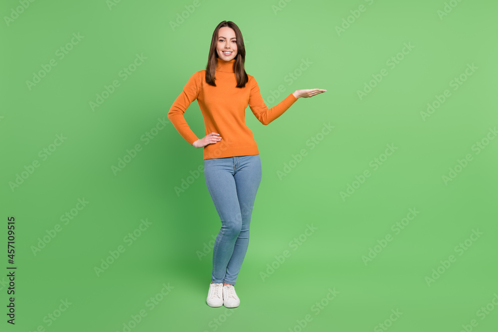 Full length body size photo girl wearing orange keeping blank space on hand isolated pastel green color background Full length body size photo girl wearing orange keeping blank space on hand isolated pastel green color background