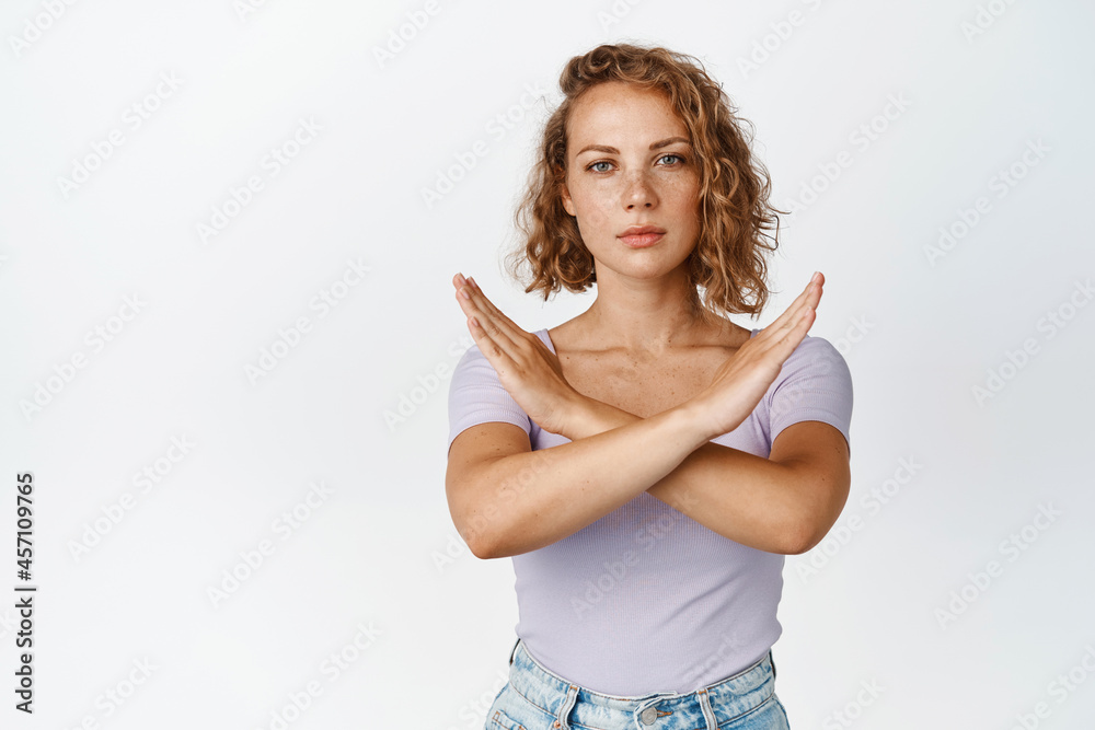 Serious young woman shows cross stop gesture, looking unamused, saying no, forbid something, standing over white background Serious young woman shows cross stop gesture, looking unamused, saying no, forbid something, standing over white background