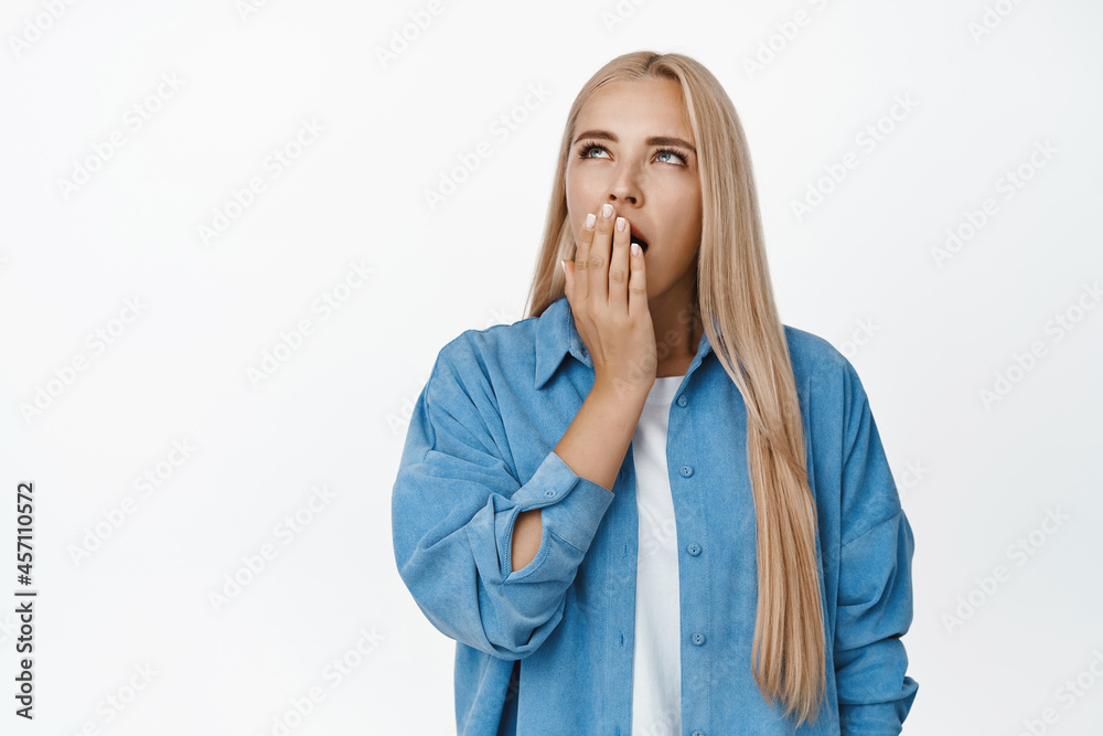 Portrait of yawning blond girl covering mouth with hand and looking up bored, standing tired against white background Portrait of yawning blond girl covering mouth with hand and looking up bored, standing tired against white background