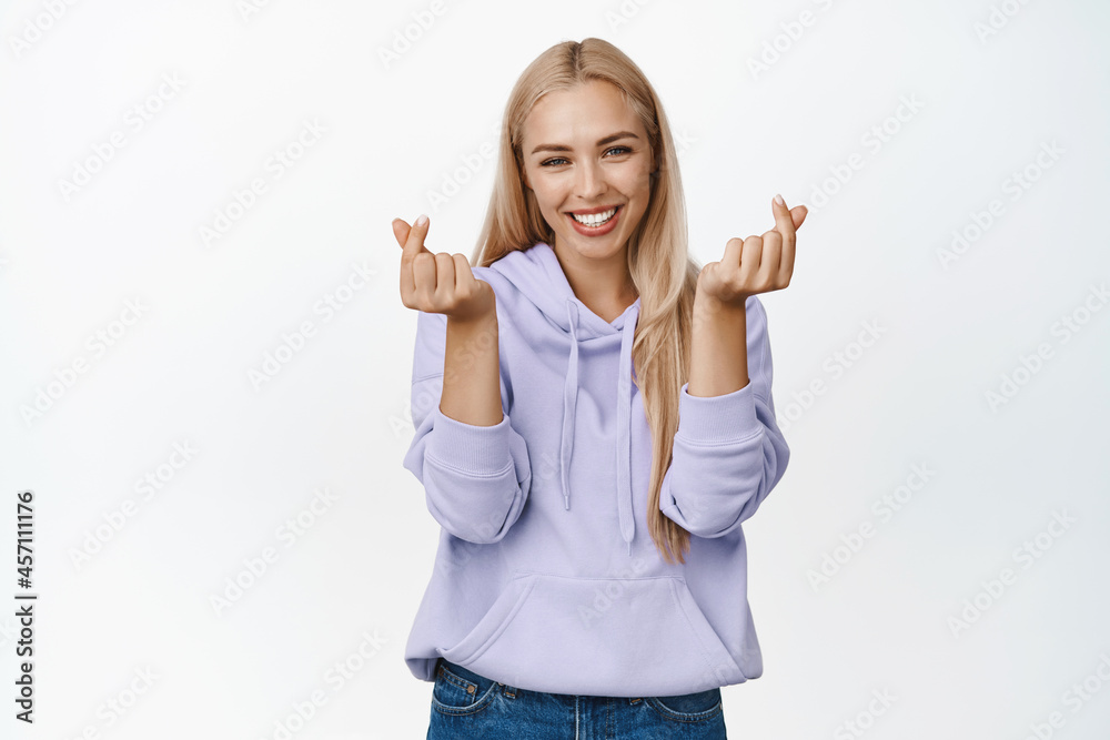 Cute blond girl in hoodie showing finger heart gesture, smiling and looking lovely at camera, standing in purple hoodie over white background Cute blond girl in hoodie showing finger heart gesture, smiling and looking lovely at camera, standing in purple hoodie over white background