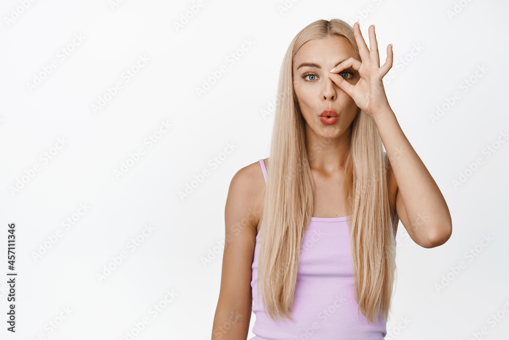 Image of blond girl looking surprised, showing okay, zero gesture over eye, curious about something, standing over white background Image of blond girl looking surprised, showing okay, zero gesture over eye, curious about something, standing over white background
