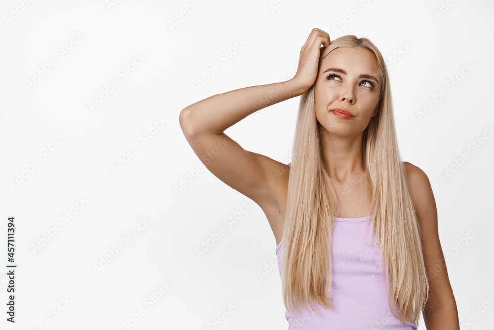 Thoughtful blond woman looking aside and scratching head, thinking, making decision, standing in pink tank top over white background
