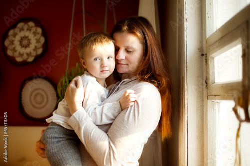 Pregnant caucasian mother with long hair with a sad baby toddler in her arms at the window in a lifestyle interior, dark brown wall, mustard wall