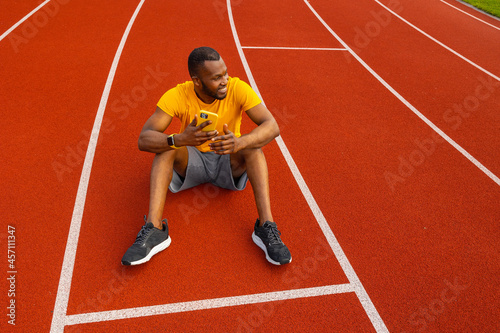 Happy attractive male athlete sitting at the stadium track, relaxing, taking a break after running. Handsome african american man using smartphone, browsing internet, smiling, workout outdoors concept