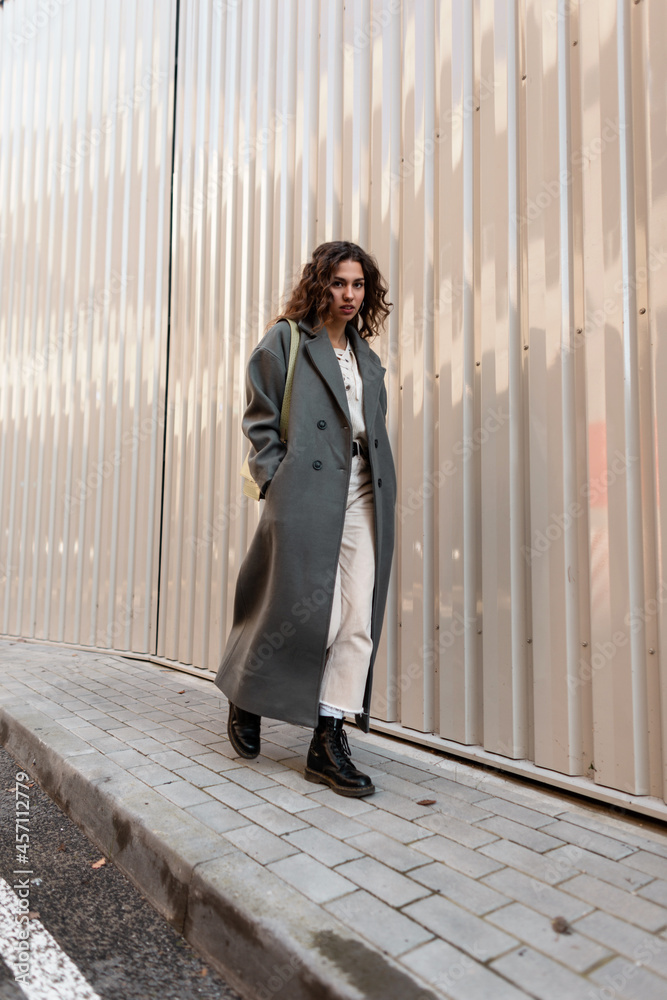 Stylish urban hipster girl with curly hair in a fashionable long green coat with a bag and boots walks on the street near a metal wall Stylish urban hipster girl with curly hair in a fashionable long green coat with a bag and boots walks on the street near a metal wall