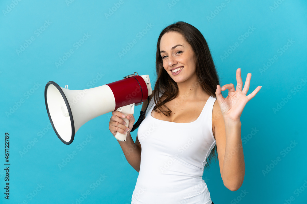 Young hispanic woman over isolated blue background holding a megaphone and showing ok sign with fingers Young hispanic woman over isolated blue background holding a megaphone and showing ok sign with fingers
