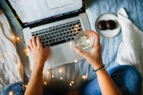 Working laptop and a cup of coffee with hands, no face. Freelancer woman working in the bedroom on the bed with a glass of wine, mock up,