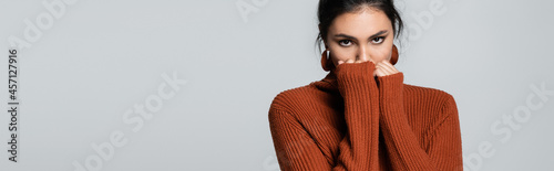 young woman in knitted sweater looking at camera while covering face isolated on grey, banner