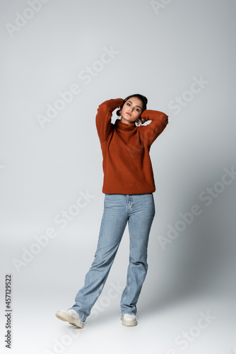 full length of young woman in sweater and denim jeans posing on grey