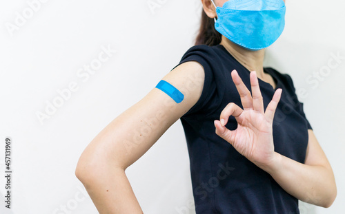 Asian woman wear face mask using adhesive bandage with blue at arm and showing O.K. gesture after injection vaccine. Covid 19 vaccine disease preparing.