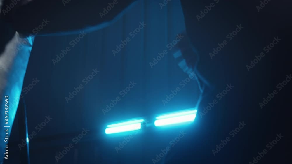 A male figure in black punching a boxing bag at a very fast pace with his fists. In the background you can see glowing blue lights in a dark room setting.
