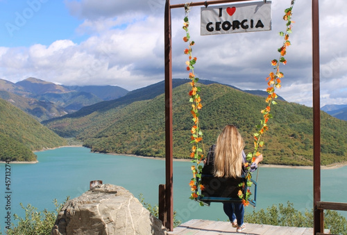 The girl rides on a swing in front of the cliff, admiring the panoramas of the mountains and the lake