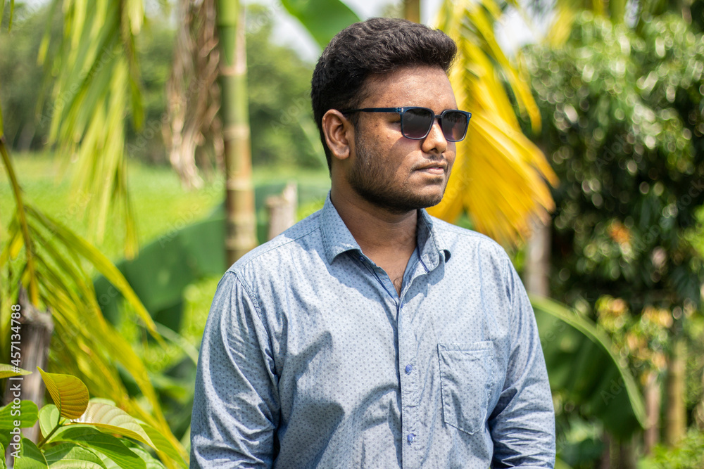 A handsome young man is standing in colorful sunglasses and a white-blue shirt, with a blurry green background behind him A handsome young man is standing in colorful sunglasses and a white-blue shirt, with a blurry green background behind him