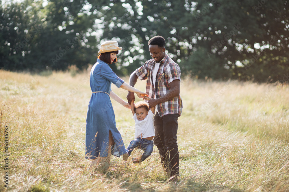 Beautiful multi ethnic parents in casual wear playing with little happy son on field. Positive family relaxing together on fresh air. Summer time and lifestyles concept.