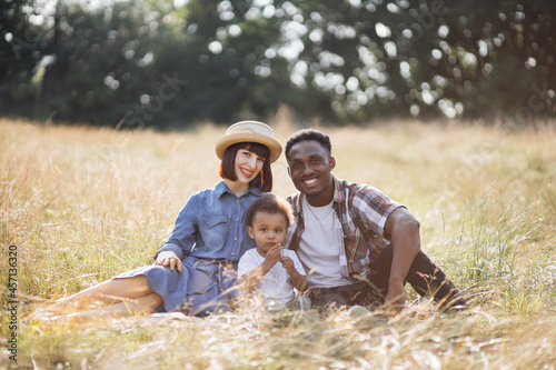 Portrait of beautiful and happy family of three having summer pinic on nature. Cute little boy sitting in hugs with multicultural parents and eating sweet grape.