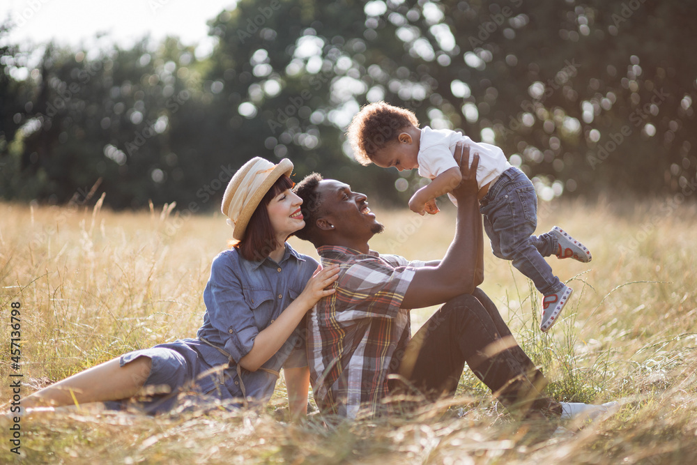 Positive multi ethnic family of three in casual wear playing together during sunny day outdoors. Caucasian woman hugging her african husband that lifting little son on hands. Positive multi ethnic family of three in casual wear playing together during sunny day outdoors. Caucasian woman hugging her african husband that lifting little son on hands.