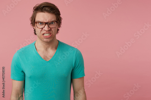 indoor shot of young brunette man, wears glasses and blue t-shirt, looks directly into camera with mad negative facial expression. Isolated over pink background © timtimphoto