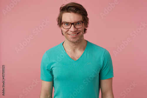 Indoor shot of young positive male, wears stylish glasses and blue t-shirt, looks directly into camera with broad smile, feels happy. Isolated over pink background © timtimphoto