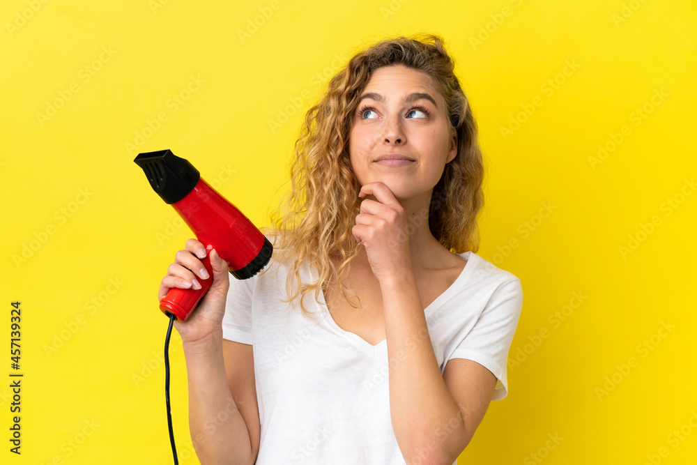 Young blonde woman holding a hairdryer isolated on yellow background and looking up Young blonde woman holding a hairdryer isolated on yellow background and looking up