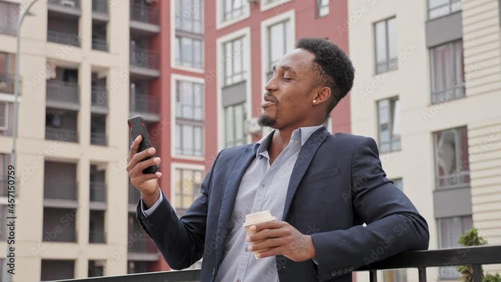 Confident black handsome man having rest dressed in formal clothing using modern smartphone for conversation drinking morning coffee to go. Video call with partners on mobile phone standing outdoors. Confident black handsome man having rest dressed in formal clothing using modern smartphone for conversation drinking morning coffee to go. Video call with partners on mobile phone standing outdoors.