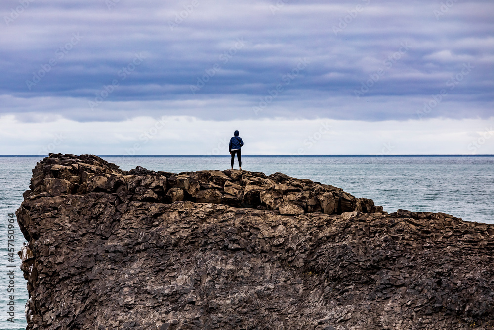 A lone traveler standing on top of a cliff facing the Atlantic ocean in Iceland. 