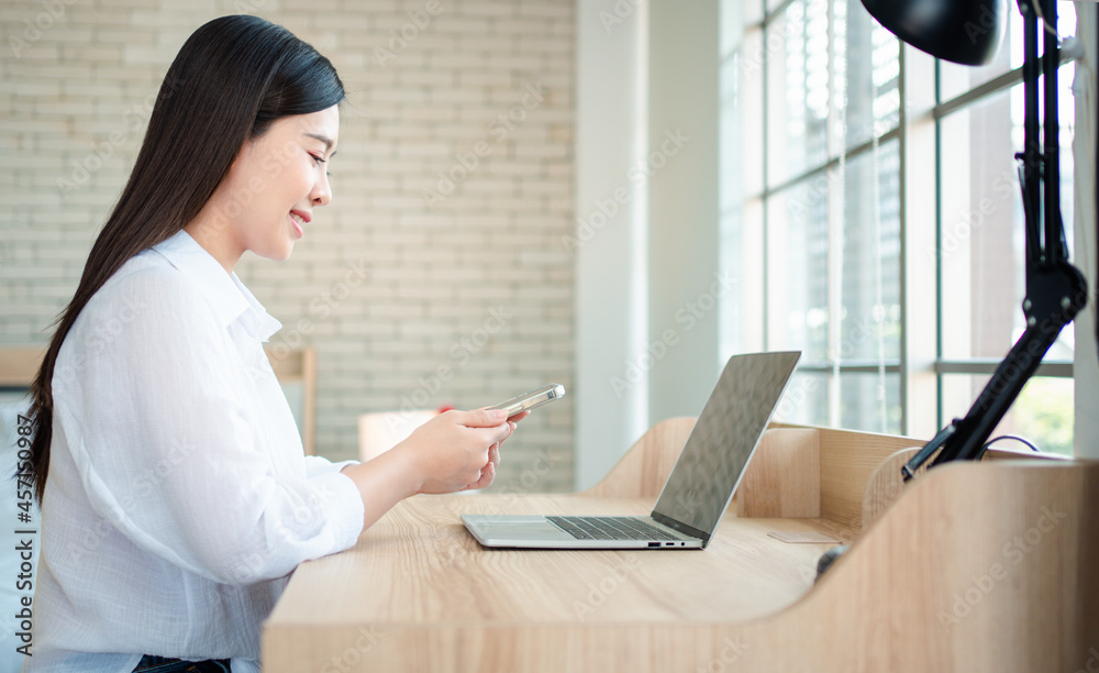 Happy beautiful asian woman working on a laptop and smartphone at the home office sitting at table. Happy female professional freelancer online using notebook pc and smartphone concept. Happy beautiful asian woman working on a laptop and smartphone at the home office sitting at table. Happy female professional freelancer online using notebook pc and smartphone concept.