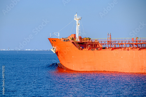 leuchtend rot oranger Frachter auf blauem stillen Meer der Bugwelle vor sich her schiebt, am Horizont die Küste mit Industrieanlagen